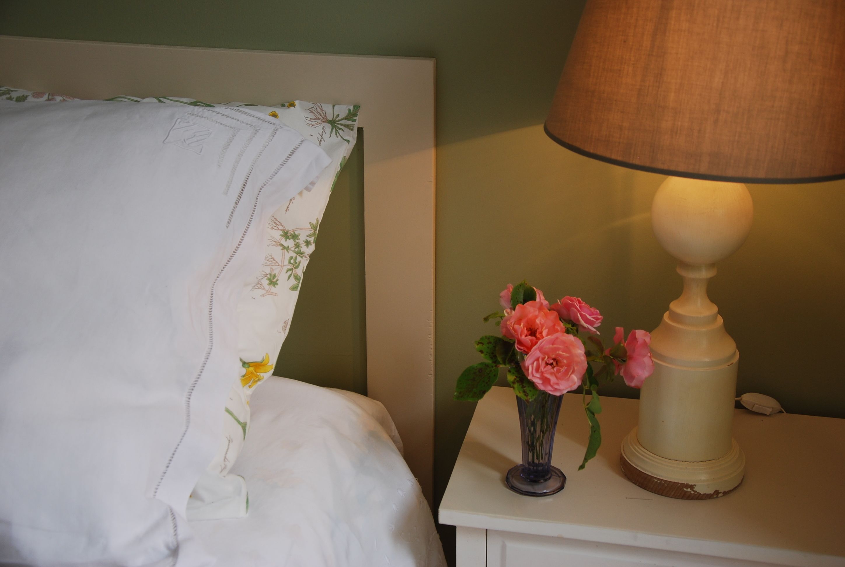 Una cama bien hecha con un cojín estampado con motivos florales y una mesita de noche con una lámpara y un florero de flores rosadas.