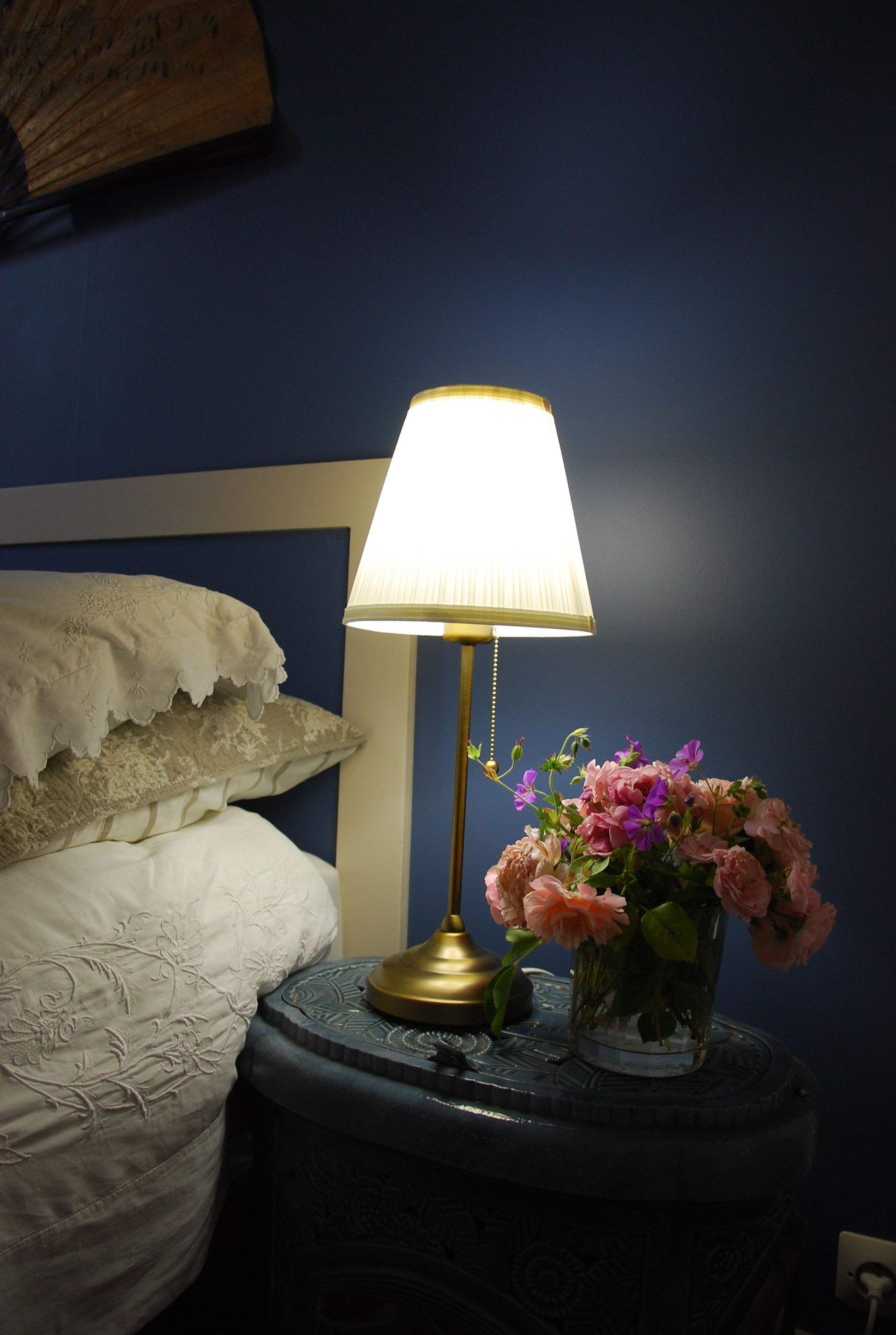 A bedside table with a lamp and a vase of flowers next to a bed with a white pillow and a decorative headboard.
