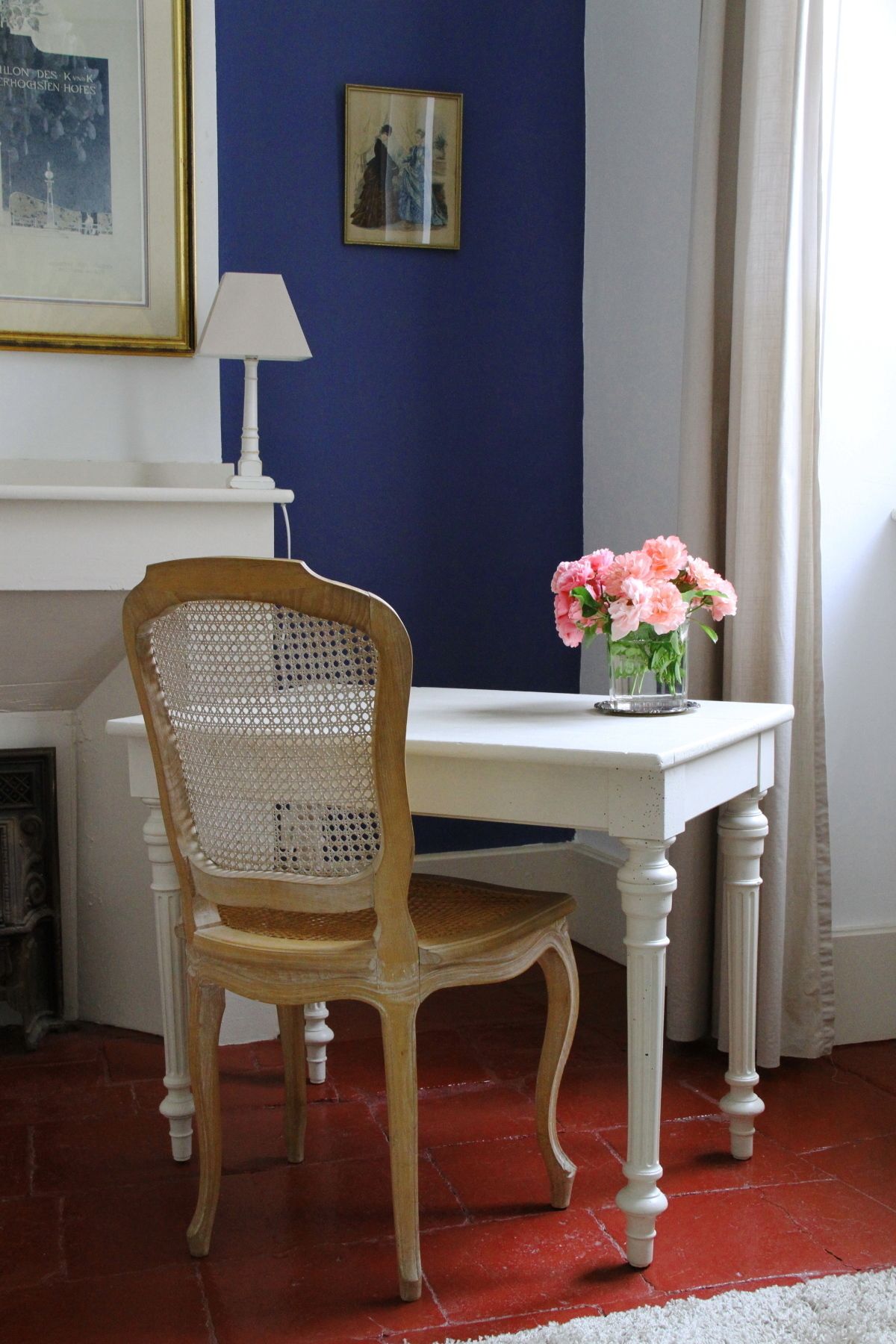 Une pièce comprenant une chaise en bois, une table blanche, un vase avec des fleurs, une lampe et un tableau encadré accroché au mur.