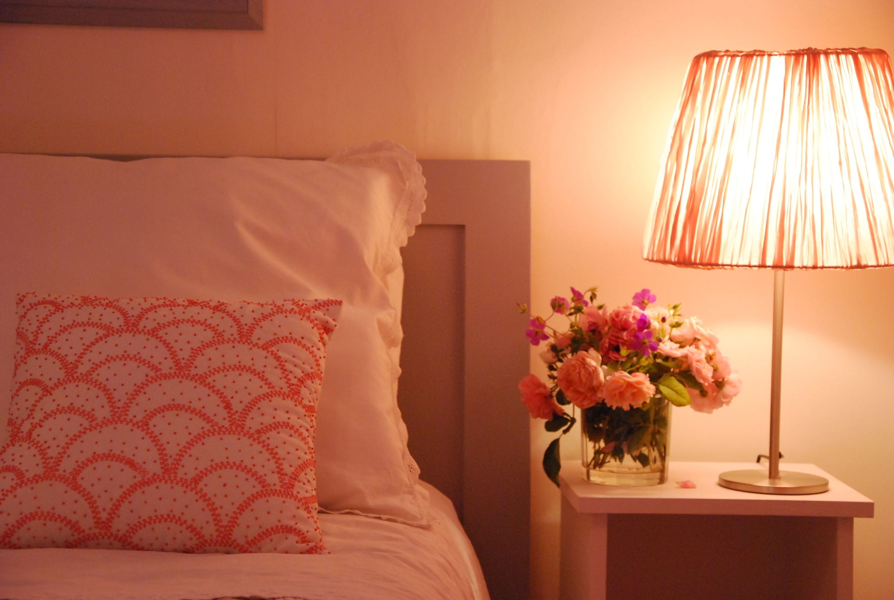 Un lit bien fait, agrémenté d’un coussin à motifs et d’une table de chevet sur laquelle se trouvent une lampe et un vase de fleurs.
