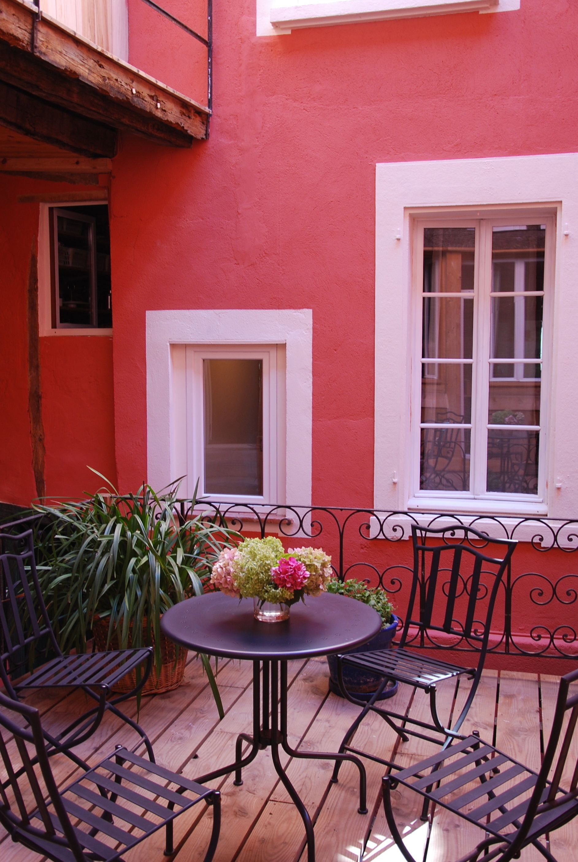 Un patio con un tavolo rotondo e quattro sedie, circondato da piante in vaso e una facciata di un edificio rossa.