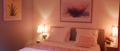 Una cama bien hecha con sábanas blancas y una colcha de flores rosa, flanqueada por dos mesitas de noche con lámparas y arreglos florales.