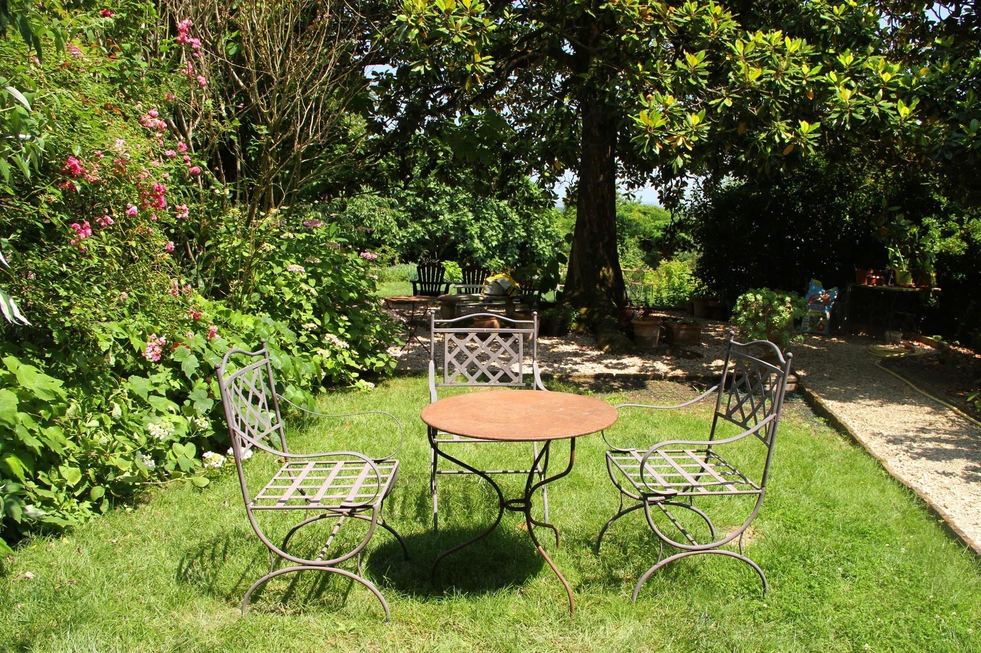 Un jardín con una mesa y sillas de metal, rodeado de follaje exuberante y plantas con flores.