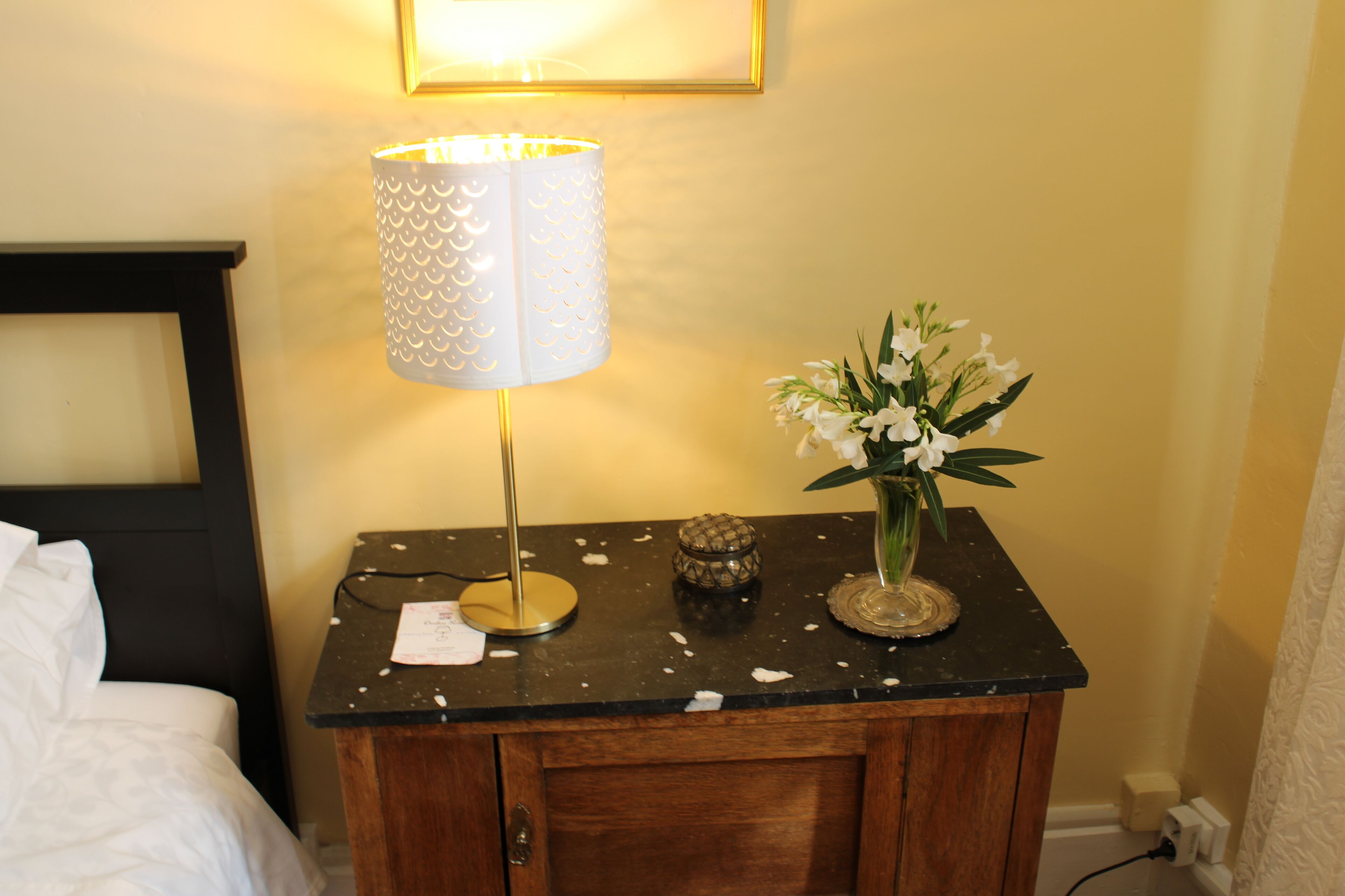 A wooden bedside table with a lamp, a vase with flowers, and a small decorative item.