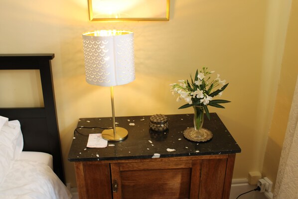 Una mesita de noche de madera con una lámpara, un florero con flores y un pequeño objeto decorativo.