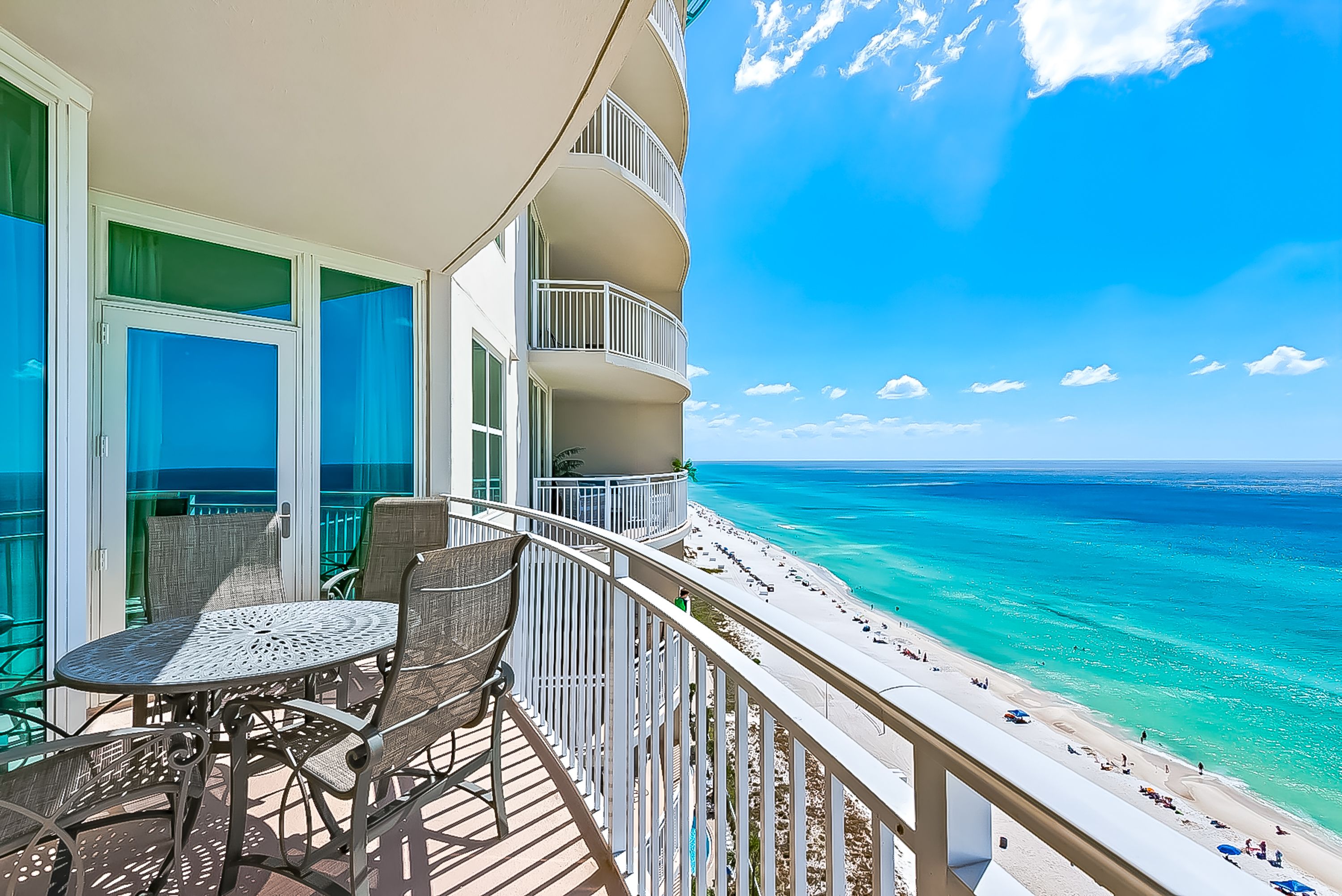 A balcony with a table and chairs, overlooking a beach with clear turquoise waters and a bright blue sky.