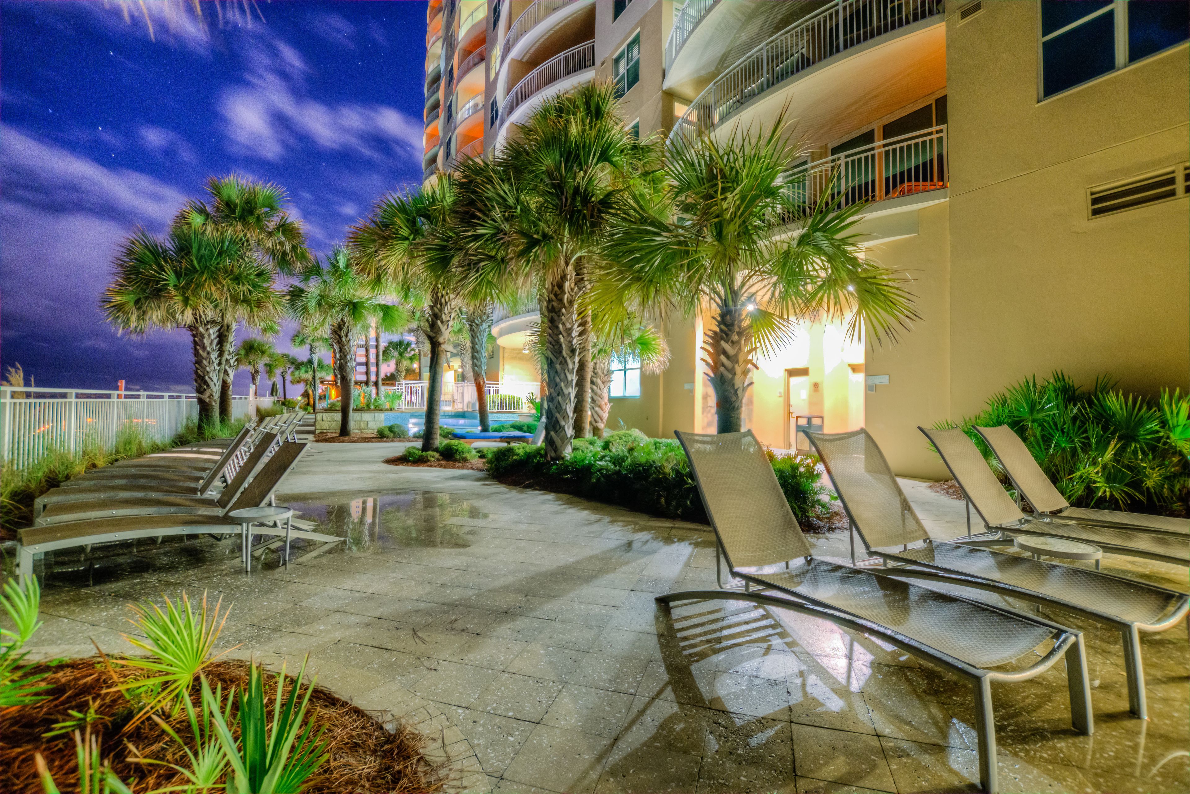 A nighttime outdoor area with lounge chairs, palm trees, and a building with balconies.