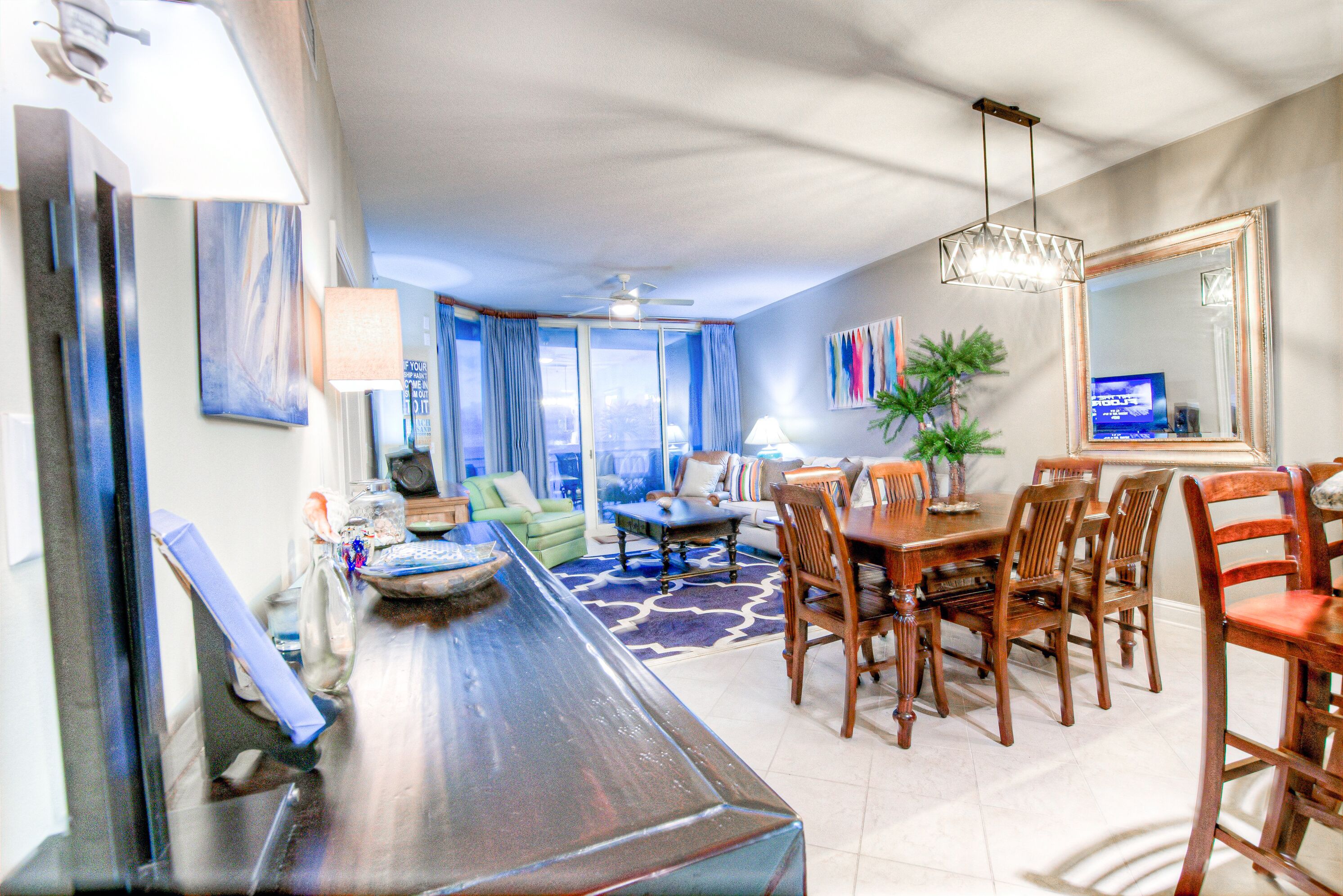 A modern living room with a dining area, featuring a long wooden table, comfortable seating, a ceiling fan, and large windows with blue curtains.