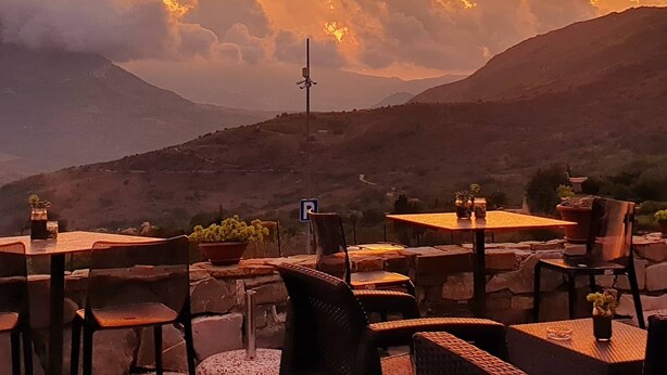 Un área de estar al aire libre con mesas y sillas, un cartel de estacionamiento y un paisaje montañoso al atardecer.