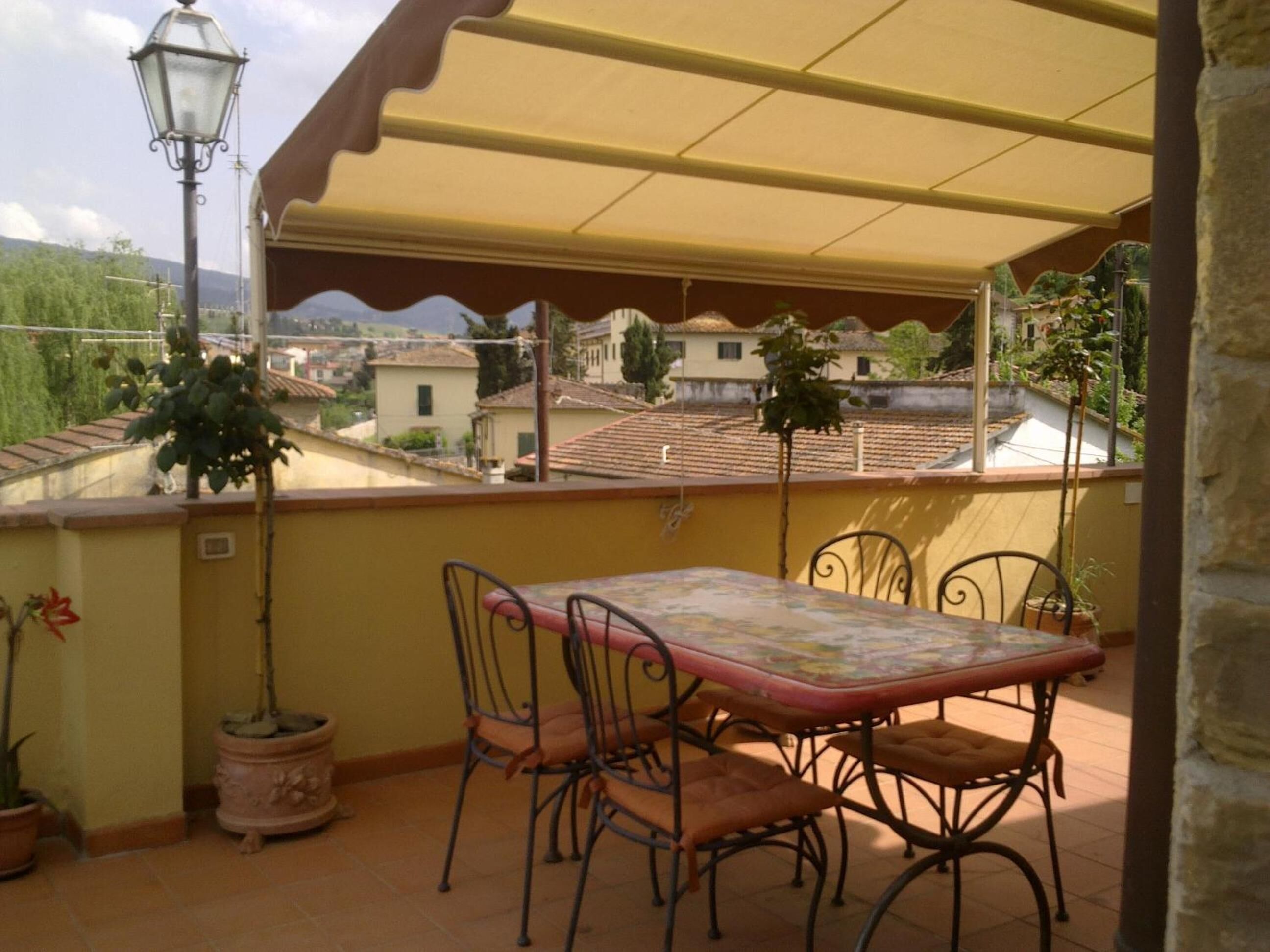 Een overdekt terras buiten met een tafel, stoelen, potplanten en uitzicht op een woonwijk.