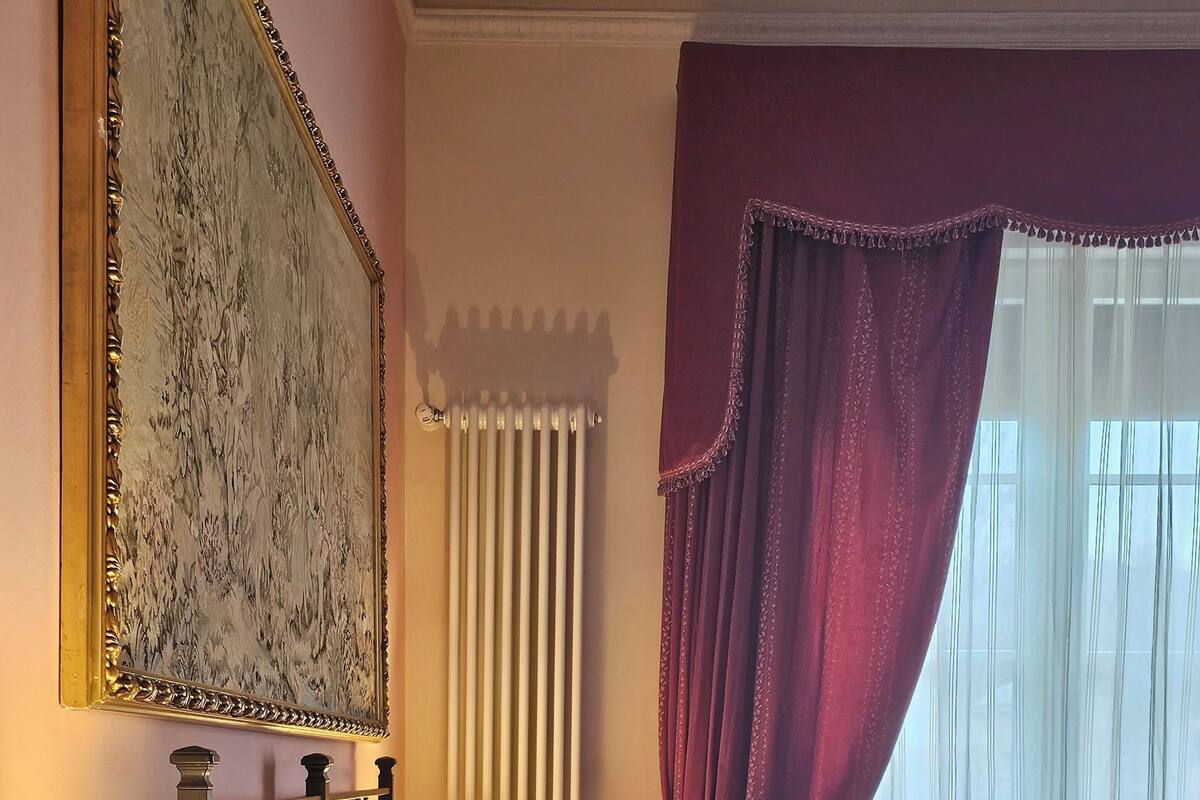 A hotel room with a bed, a nightstand with a lamp, a radiator, and a window with curtains.