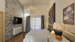 A bedroom with a stone wall, a TV, a chandelier, a bed, a nightstand, and a window with curtains.