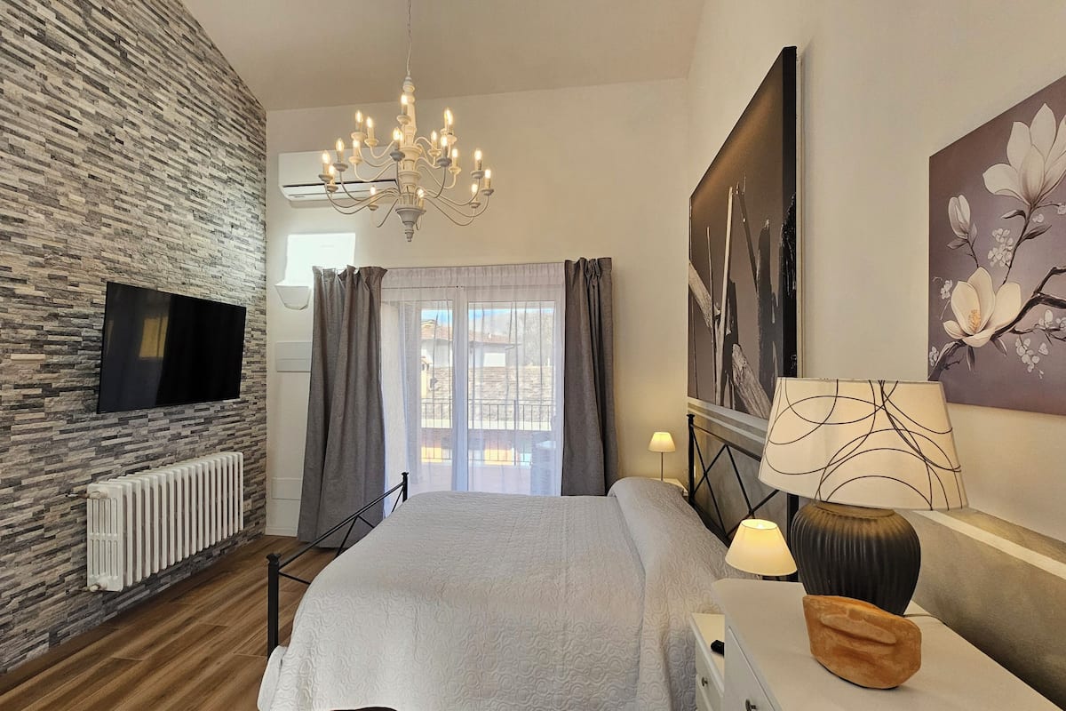 A bedroom with a stone wall, a TV, a chandelier, a bed, a nightstand, and a window with curtains.