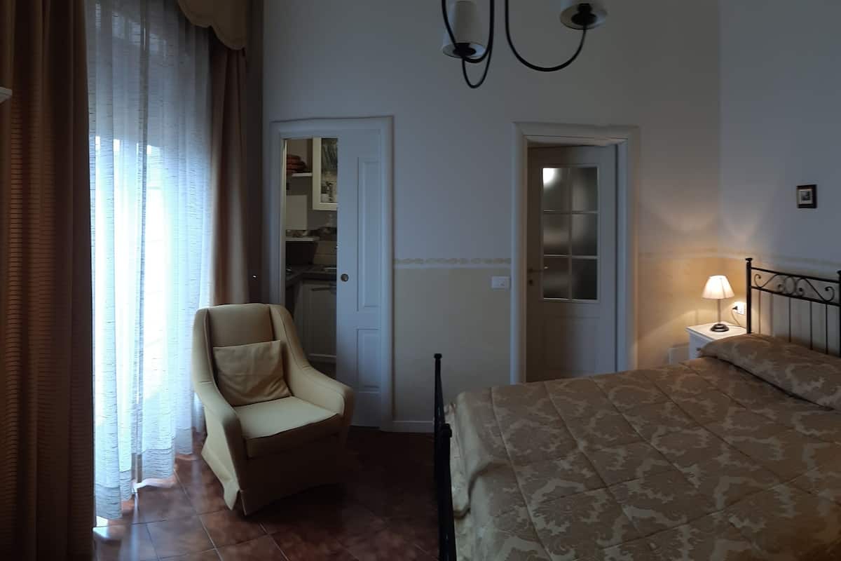 A bedroom with a bed, a chair, a nightstand with a lamp, and a door leading to another room.