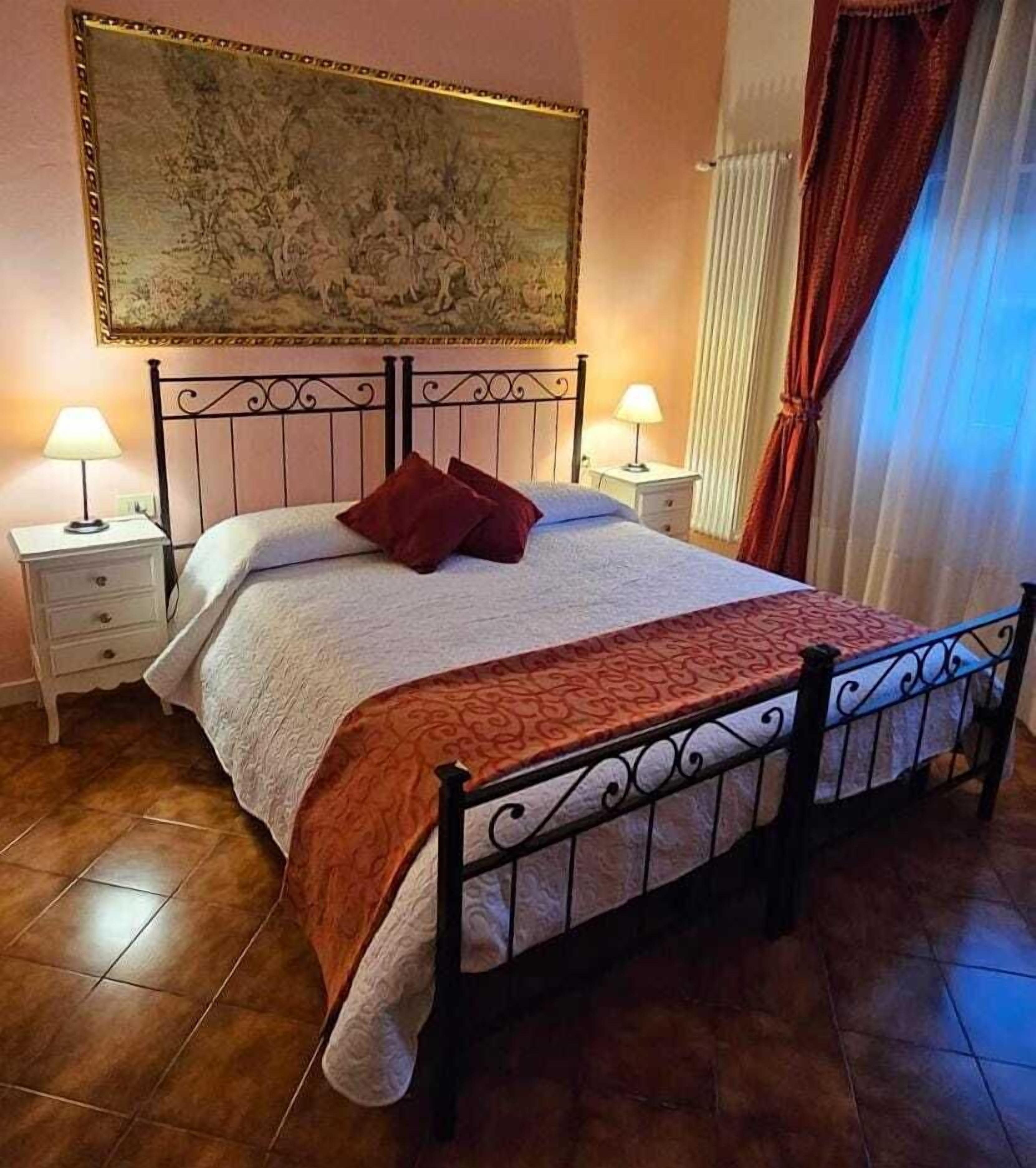 A bedroom with a bed, two bedside tables, a nightstand, a large painting on the wall, and a window with curtains.