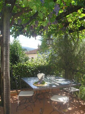 Un área de comedor al aire libre con una mesa y sillas, rodeada de follaje y con vista a un edificio al fondo.