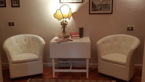 A room with two white armchairs, a small white table with a lamp, a rug, and framed pictures on the wall.