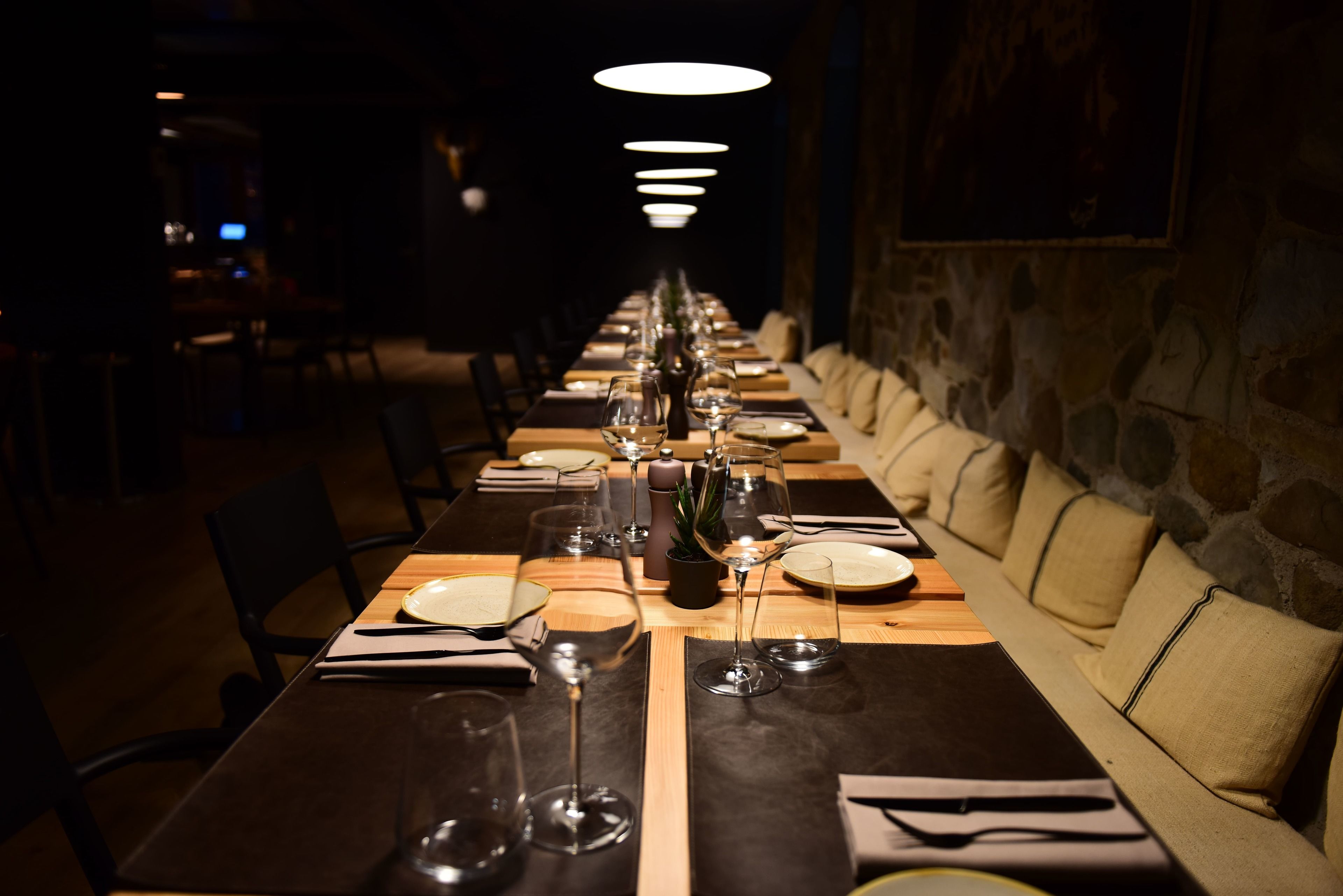 Une longue table à manger, dressée avec des assiettes, des verres et des couverts, bordée de sièges rembourrés et d’un mur en pierre.