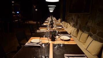 Une longue table à manger, dressée avec des assiettes, des verres et des couverts, bordée de sièges rembourrés et d’un mur en pierre.