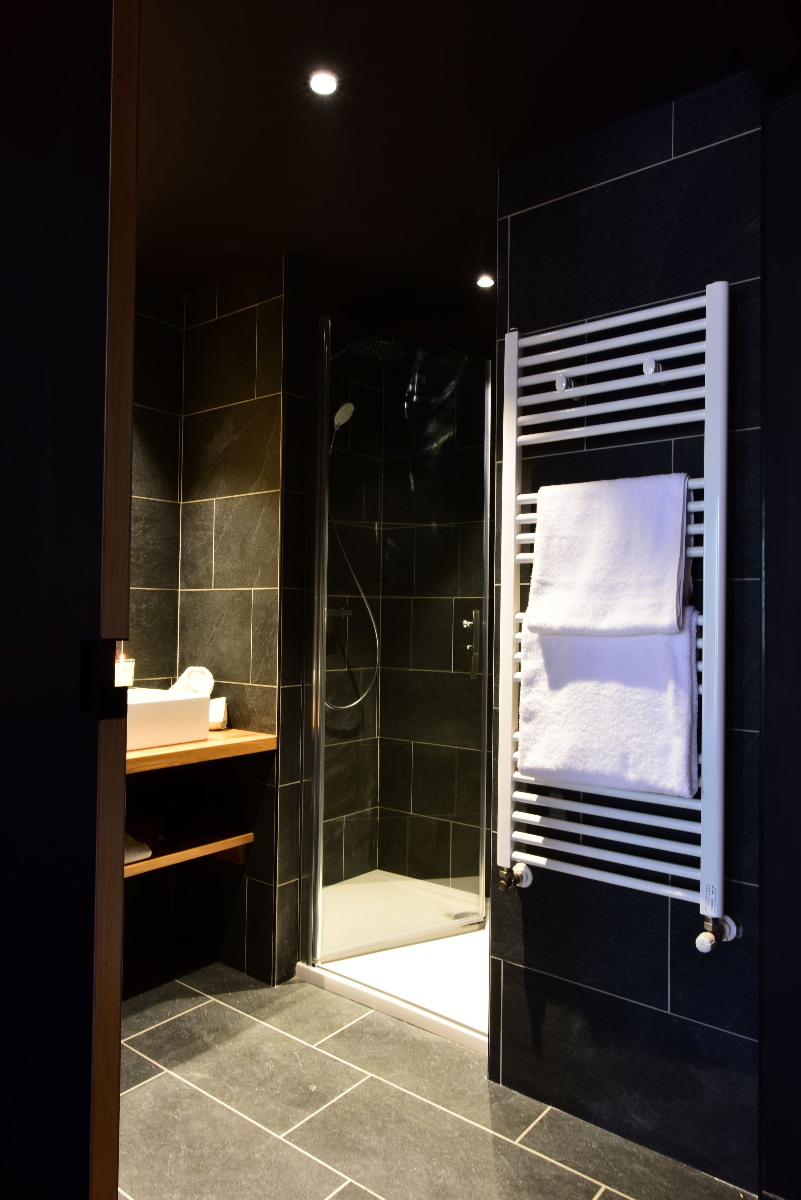 A modern bathroom with dark tiles, a glass shower enclosure, and a white towel rack.