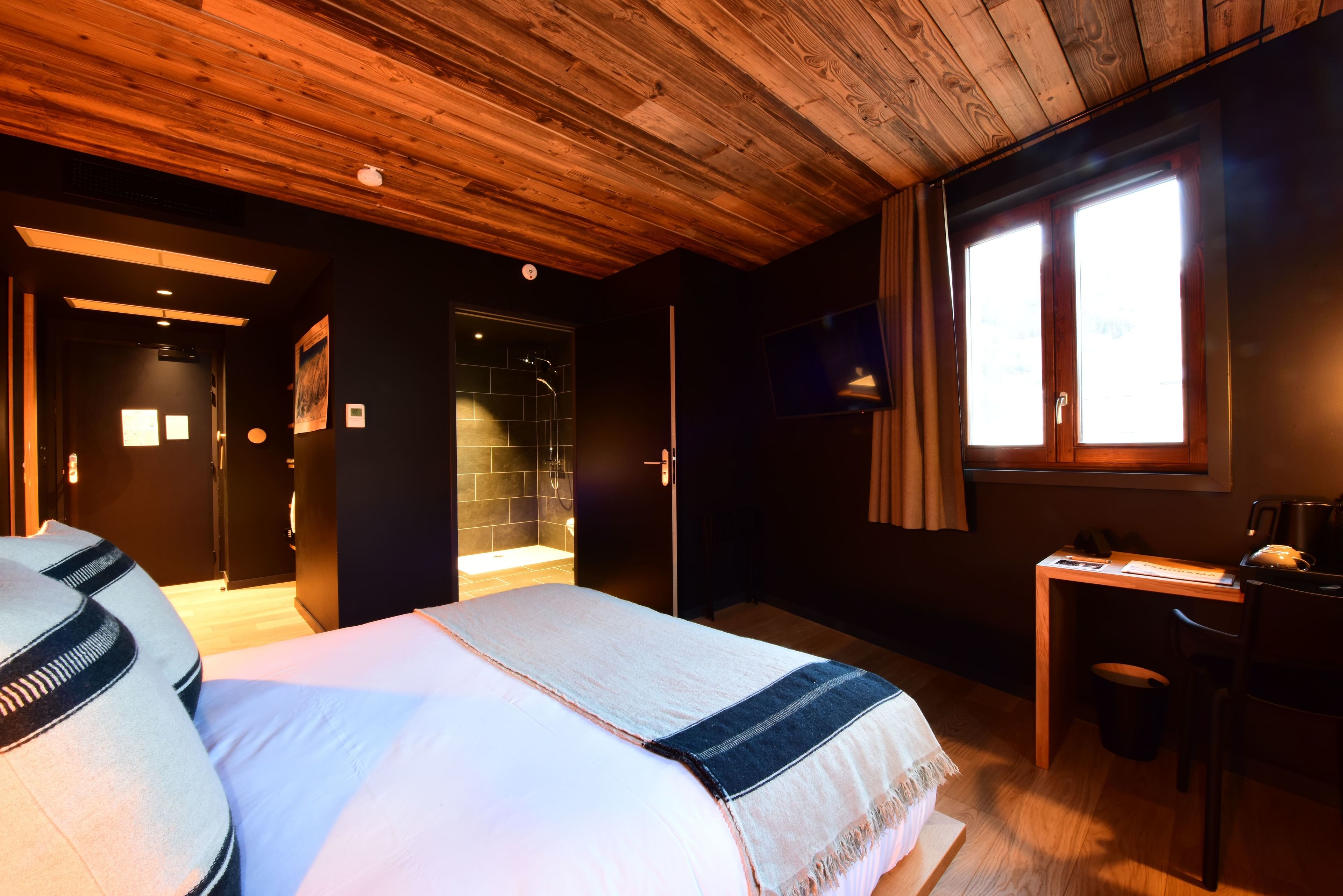 A bedroom with a wooden ceiling, a bed with a white and blue bedspread, a desk with a chair, and a window with curtains.