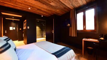 A bedroom with a wooden ceiling, a bed with a white and blue bedspread, a desk with a chair, and a window with curtains.