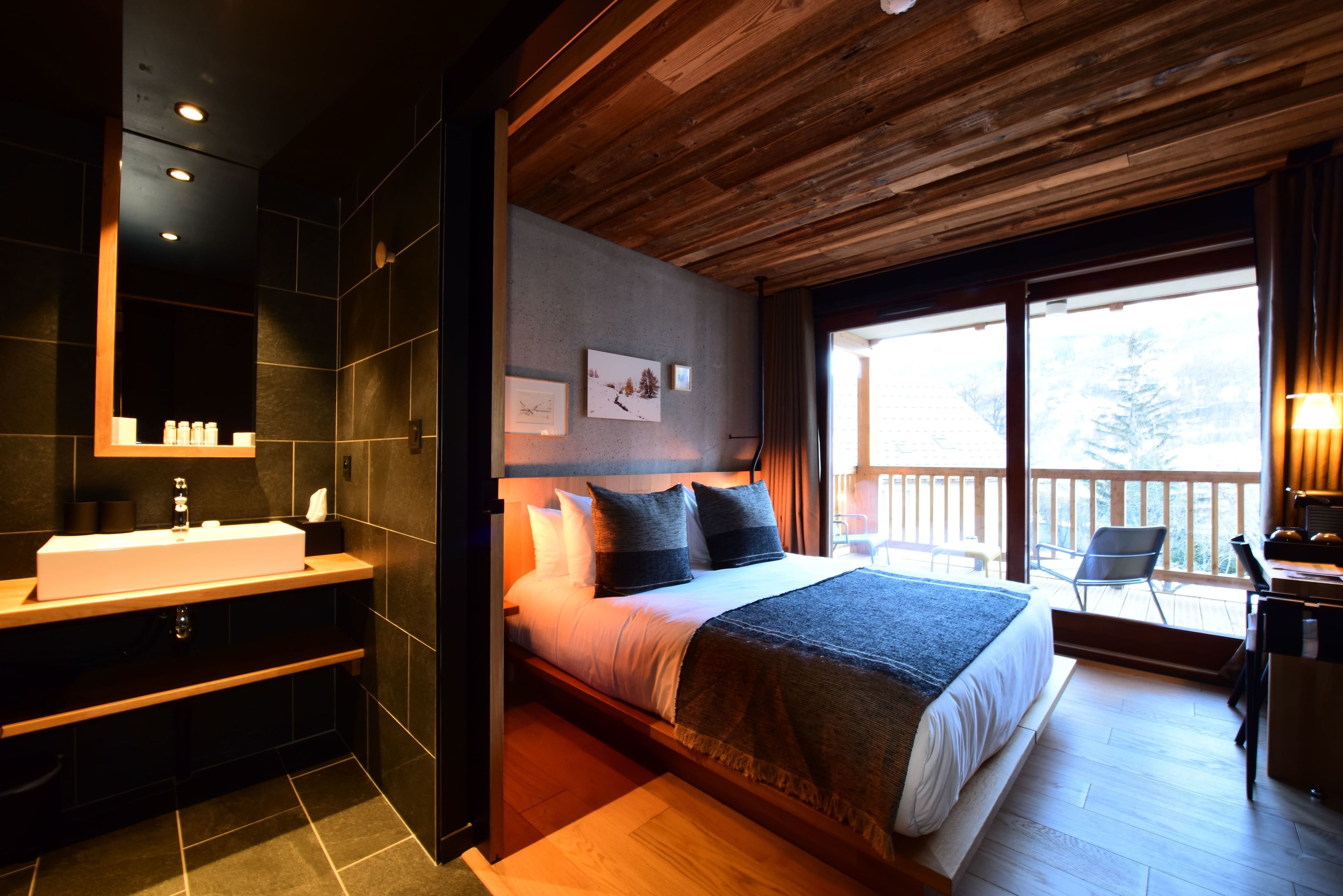 Une chambre moderne avec un grand lit, un plafond en bois, une salle de bain avec un lavabo et un balcon avec vue.