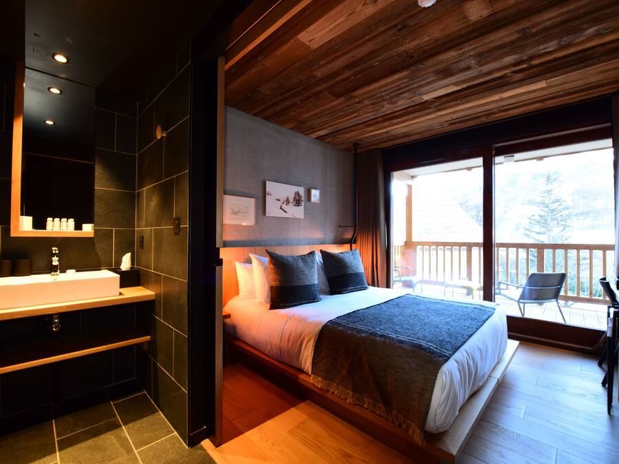 A modern bedroom with a large bed, a wooden ceiling, a bathroom with a sink, and a balcony with a view.