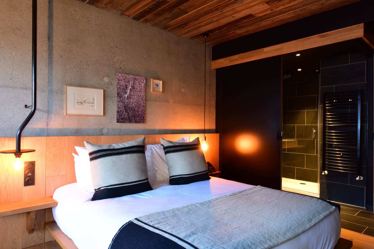 Quarto moderno com teto de madeira, uma cama grande com roupa de cama branca e duas almofadas, uma mesa de cabeceira com abajur e uma obra de arte pendurada na parede.
