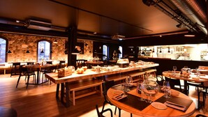 Un restaurante con varias mesas y sillas, paredes de piedra y ventanas amplias.