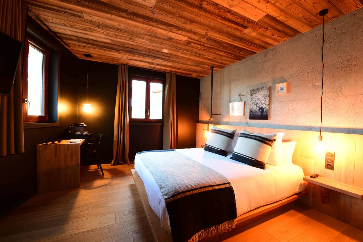 Quarto com teto de madeira, cama com roupa de cama branca e almofadas preto e branco, escrivaninha com cadeira e mesa de cabeceira com abajur.