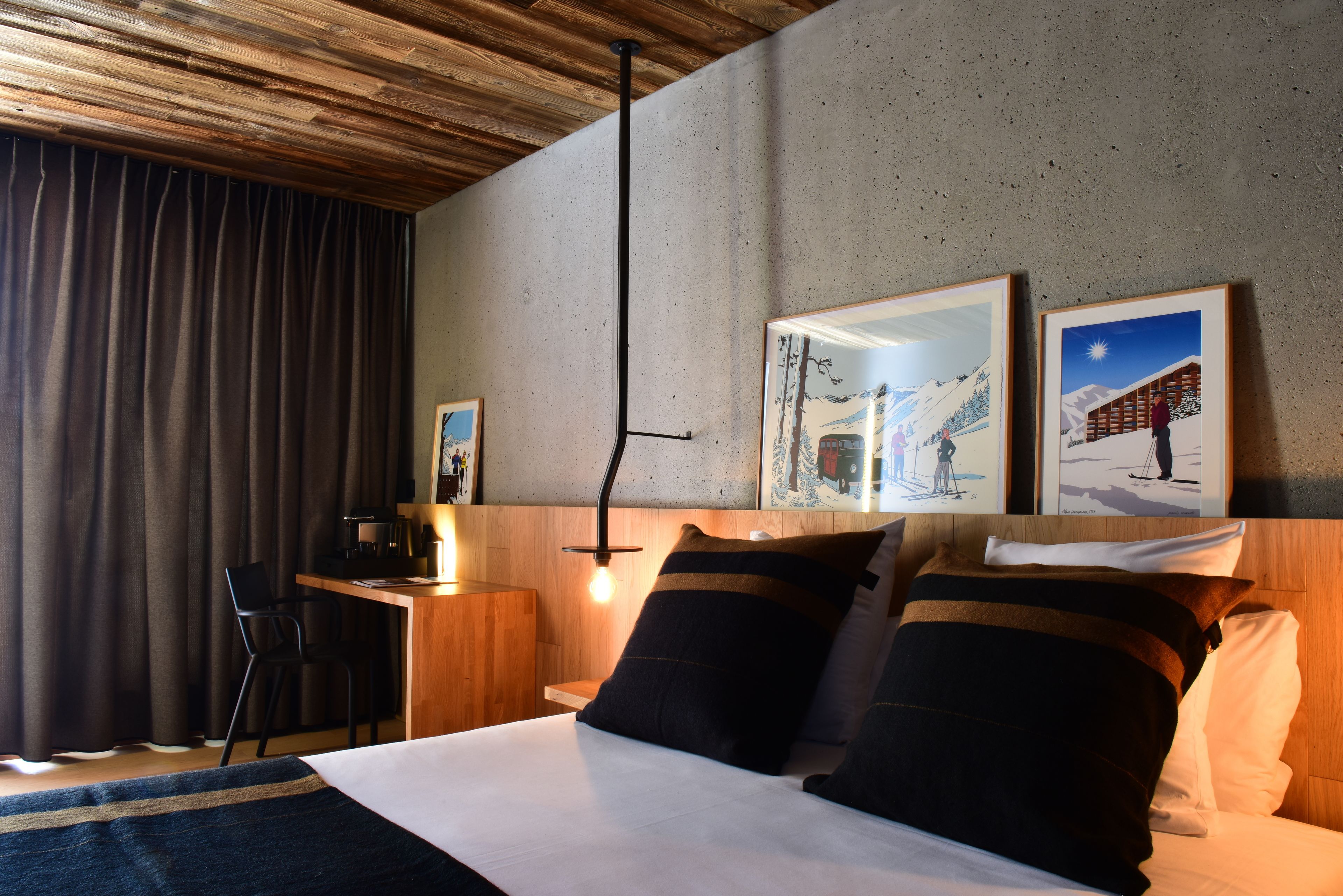 A hotel room with a wooden ceiling, a desk with a chair, a bed with two pillows, and two framed pictures on the wall.