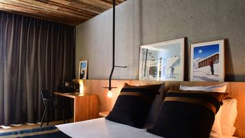 A hotel room with a wooden ceiling, a desk with a chair, a bed with two pillows, and two framed pictures on the wall.