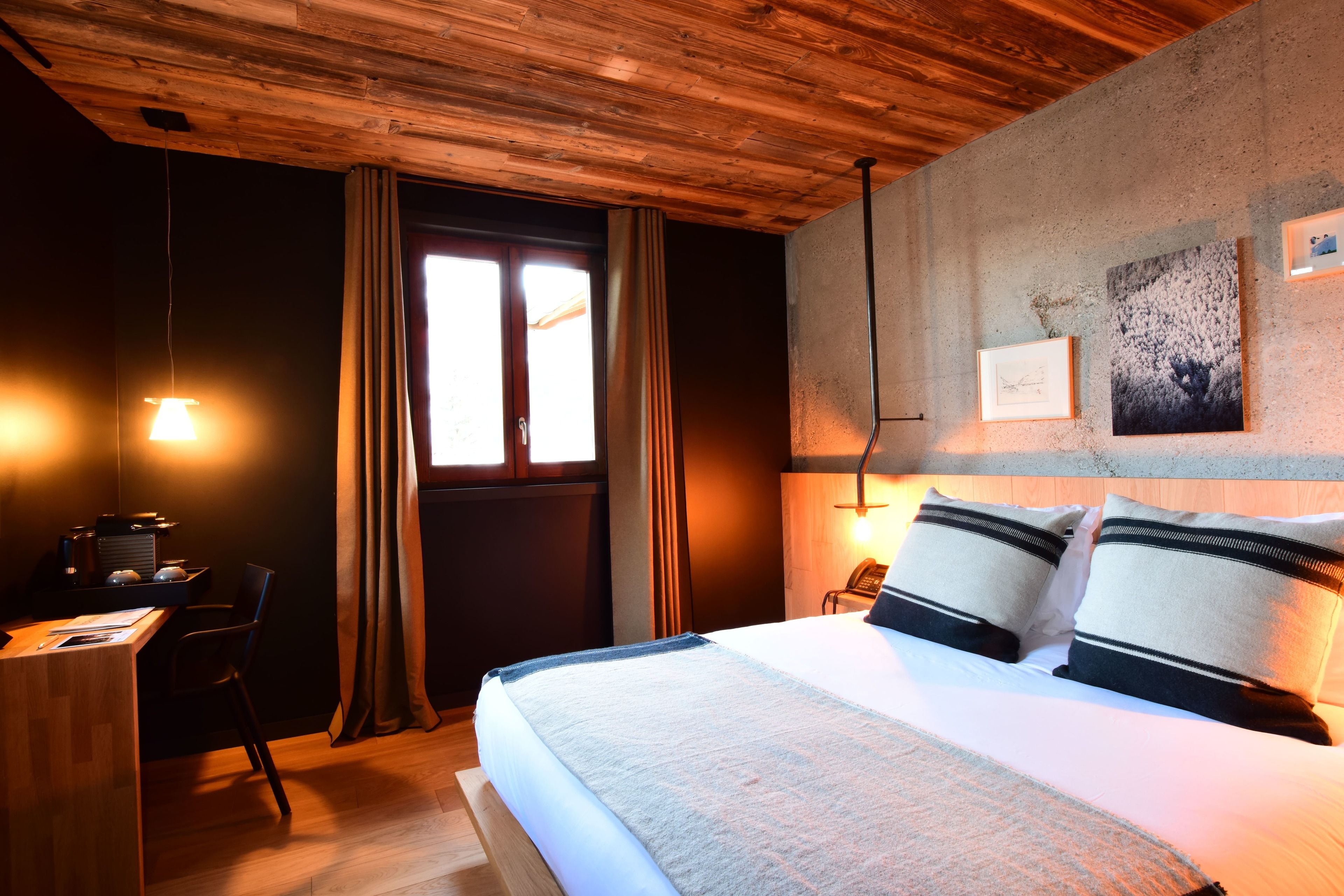 Habitación de hotel con techo de madera, cama con almohadas, escritorio con silla y ventana con cortinas.