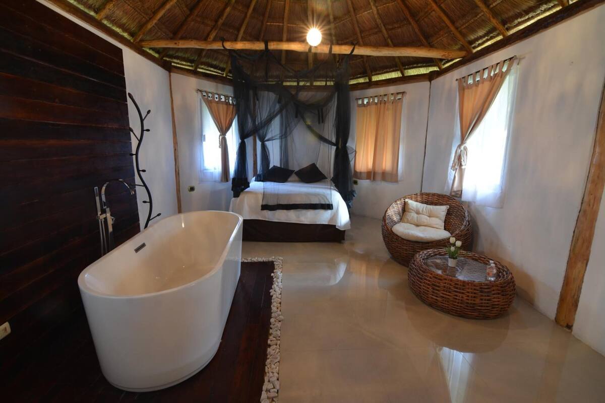 Una habitación con una bañera grande, una cama con ropa de cama negra, muebles de mimbre y un techo de paja.
