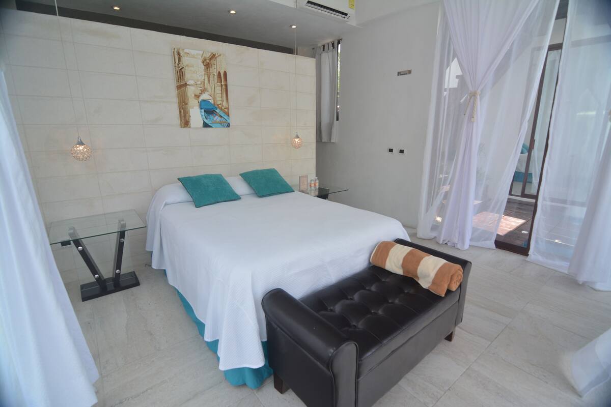 Un dormitorio moderno con cama, un banco, una mesita de vidrio y un cuadro en la pared.