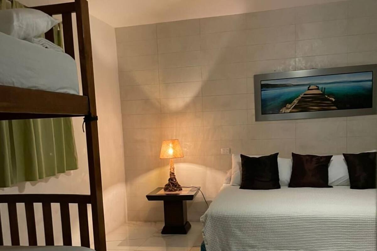 Una habitación de hotel con literas, una cama individual, una mesita de noche con lámpara, un ventilador de techo y un cuadro en la pared.