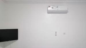 Un salon avec un canapé bleu, deux coussins à motifs, un téléviseur à écran plat fixé au mur et un appareil de climatisation placé au-dessus du canapé.