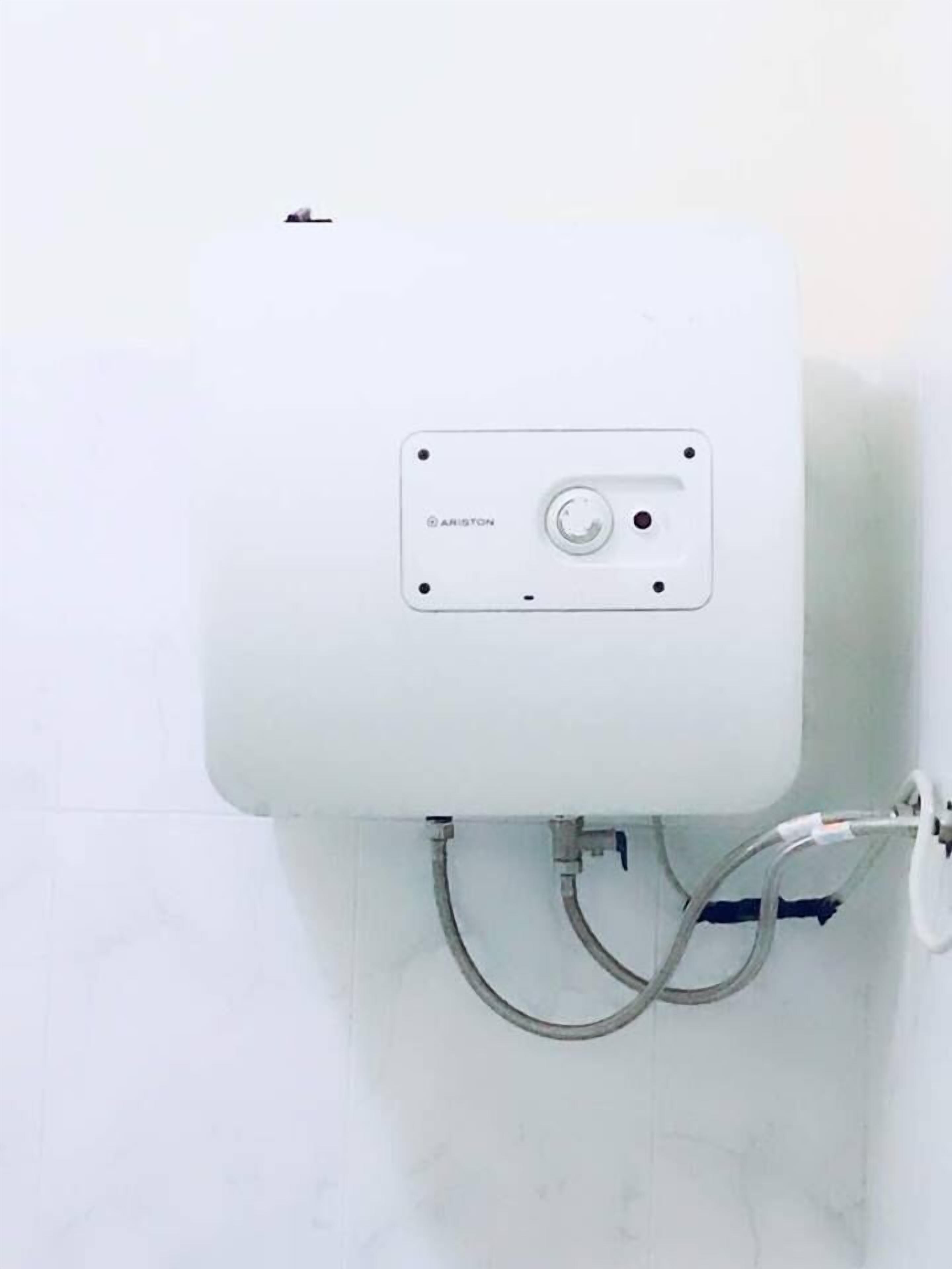 Calentador de agua blanco instalado en una pared de azulejos, con tuberías visibles y un panel de control con la etiqueta 'ARISTON'.