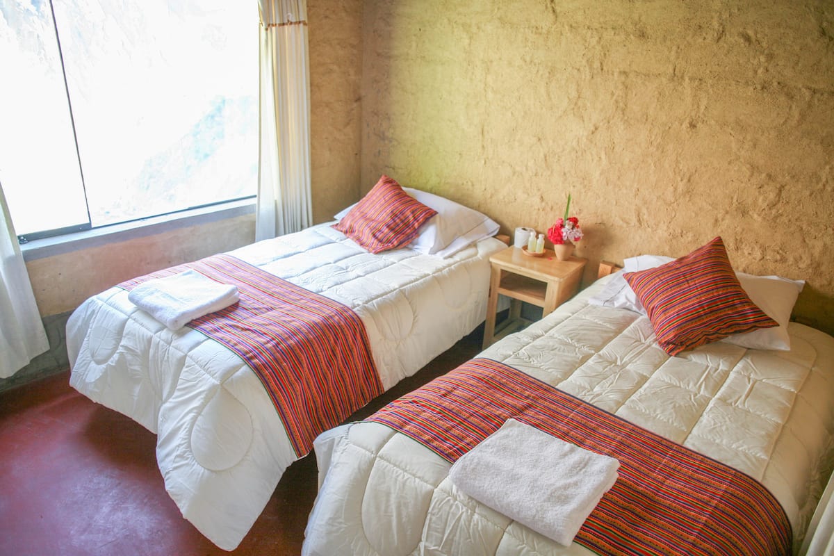 Una habitación con dos camas, cada una con ropa de cama blanca y mantas a rayas rojas. Hay una mesita de noche de madera con un florero y una botella de loción. La habitación tiene una ventana con cortinas y una pared con textura.