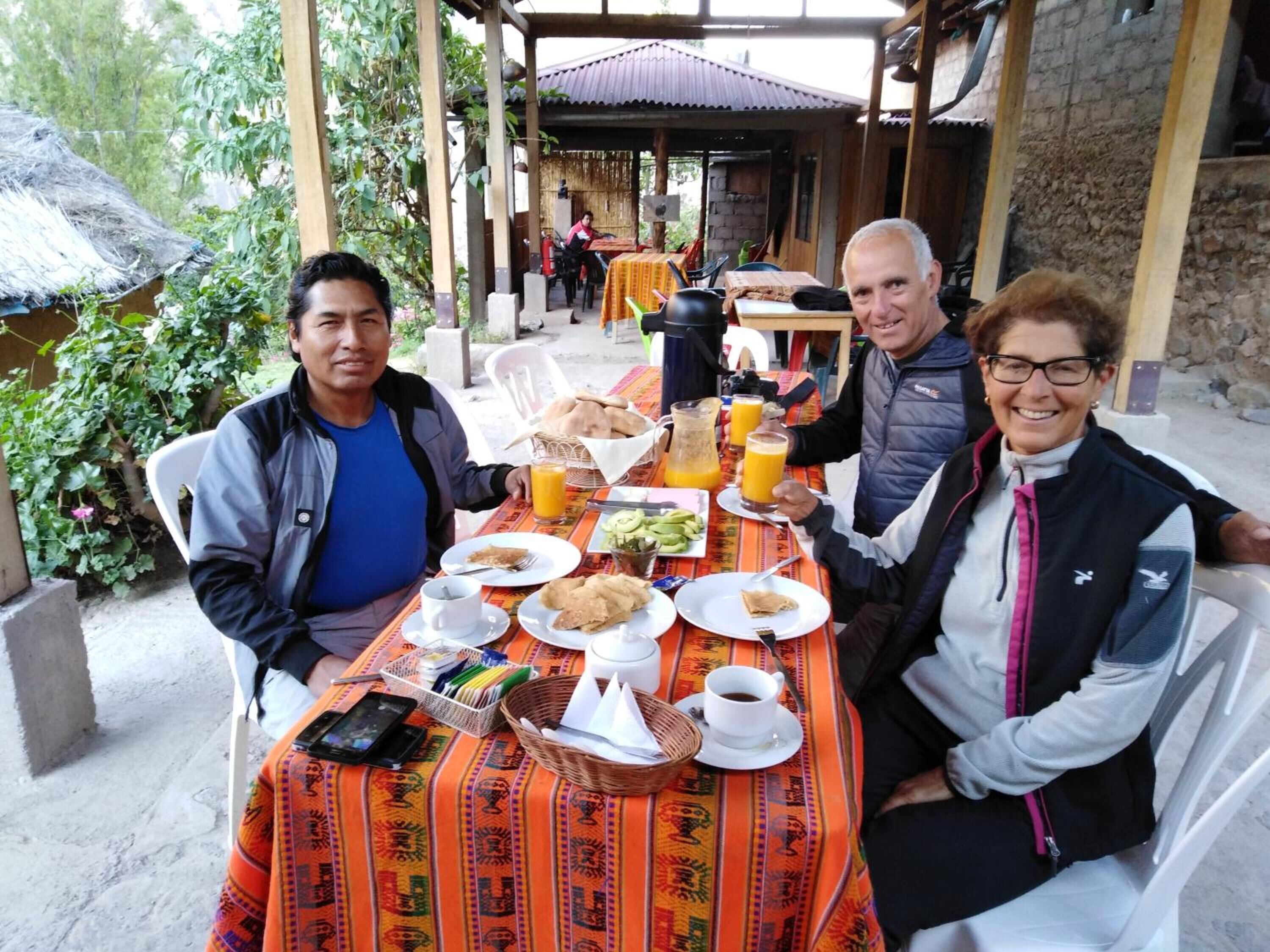 Un groupe de personnes est assis à une table en plein air, recouverte d’une nappe colorée. L’ensemble est entouré de verdure, et un bâtiment rustique se dresse en arrière-plan.