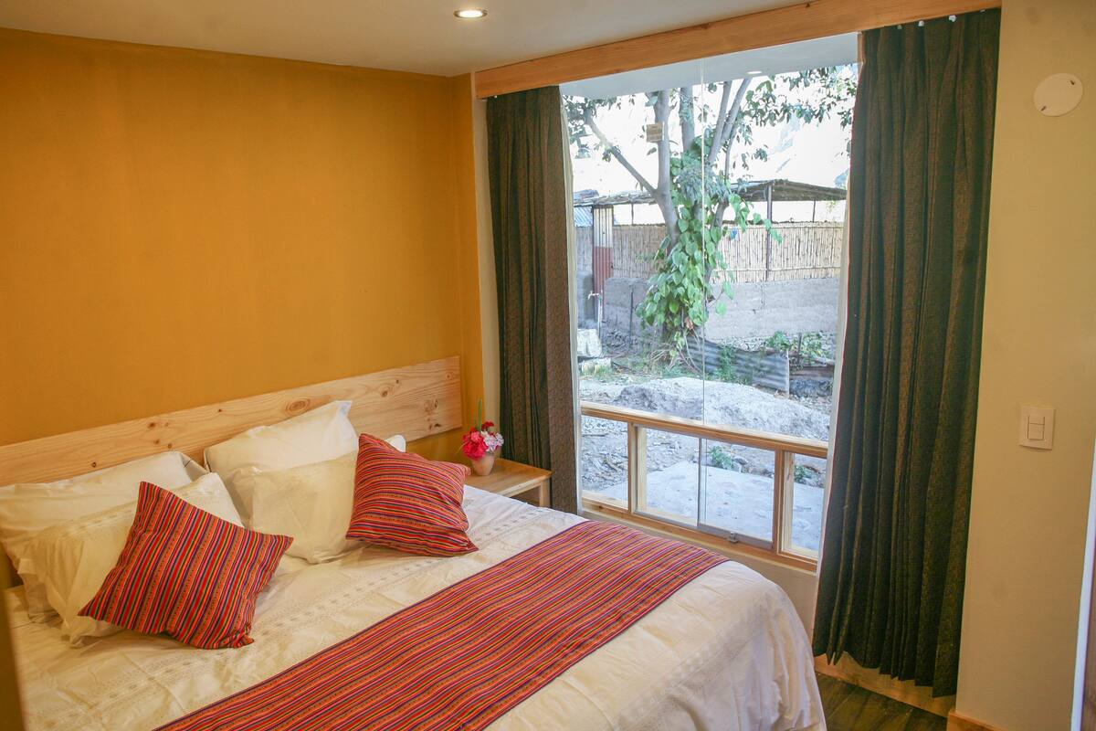 Un dormitorio con cama, almohadas, una mesita de noche y una ventana con cortinas.
