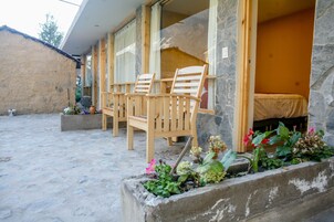 Un edificio de paredes de piedra con un banco y sillas de madera afuera, una cama adentro y plantas en macetas.