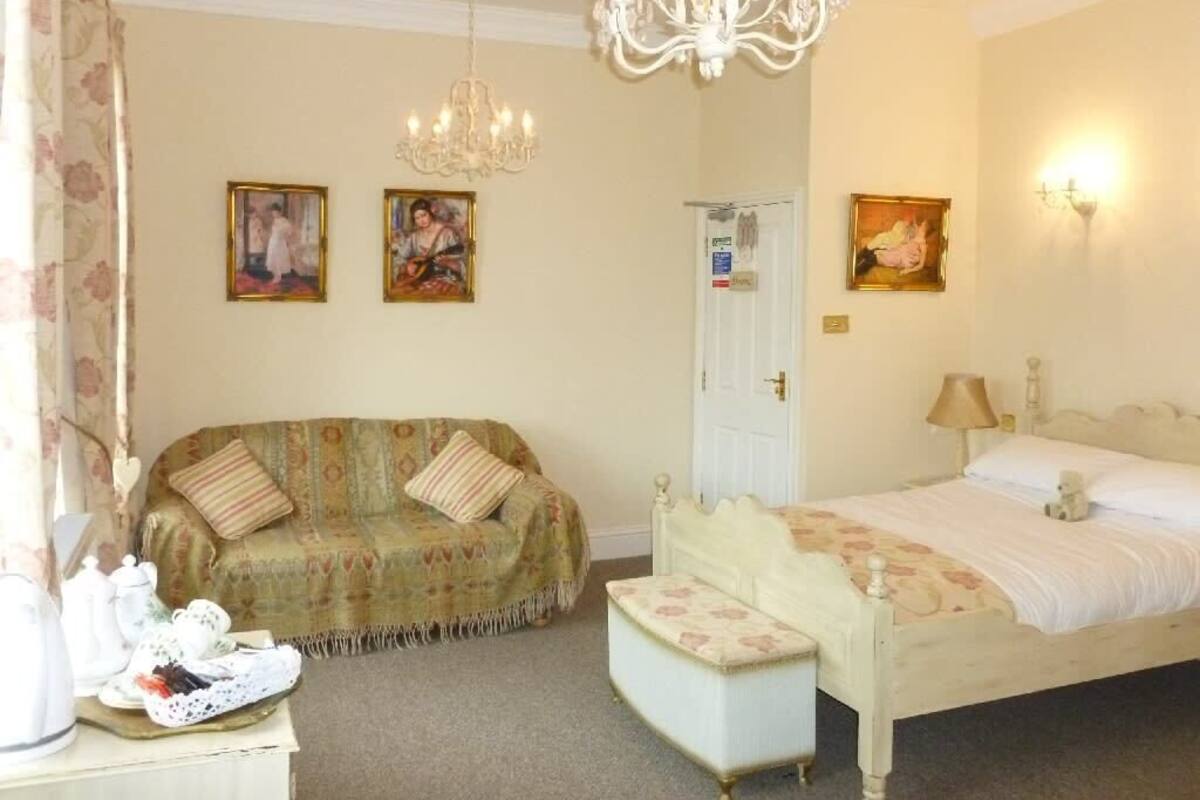 A bedroom with a bed, a bench, a sofa, a chandelier, and wall-mounted lights.