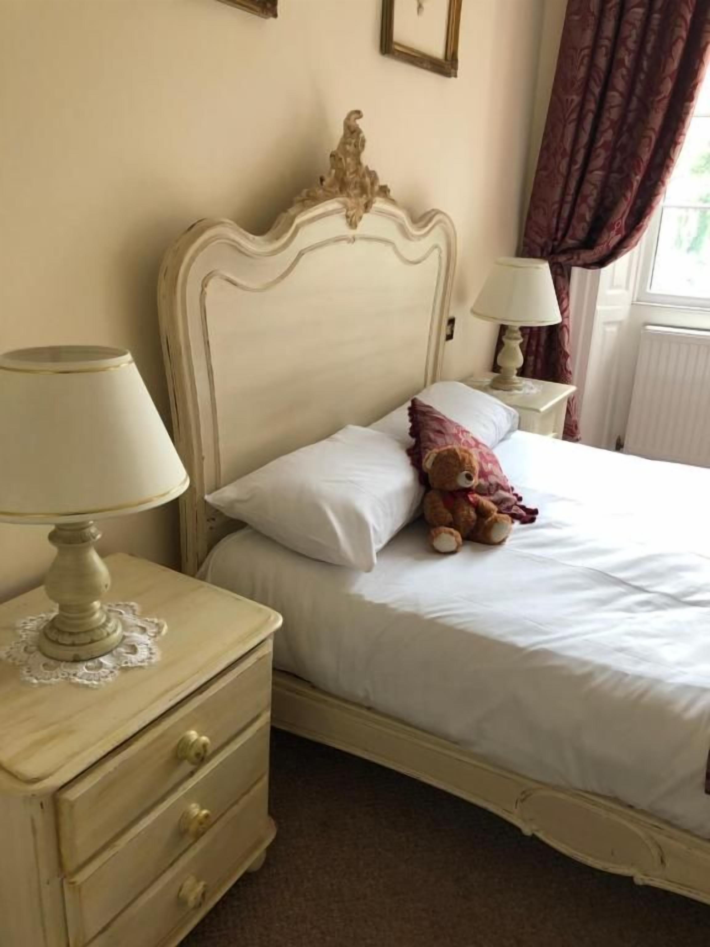 Une chambre à coucher avec un lit, une table de chevet avec une lampe, un ours en peluche et une fenêtre avec des rideaux.