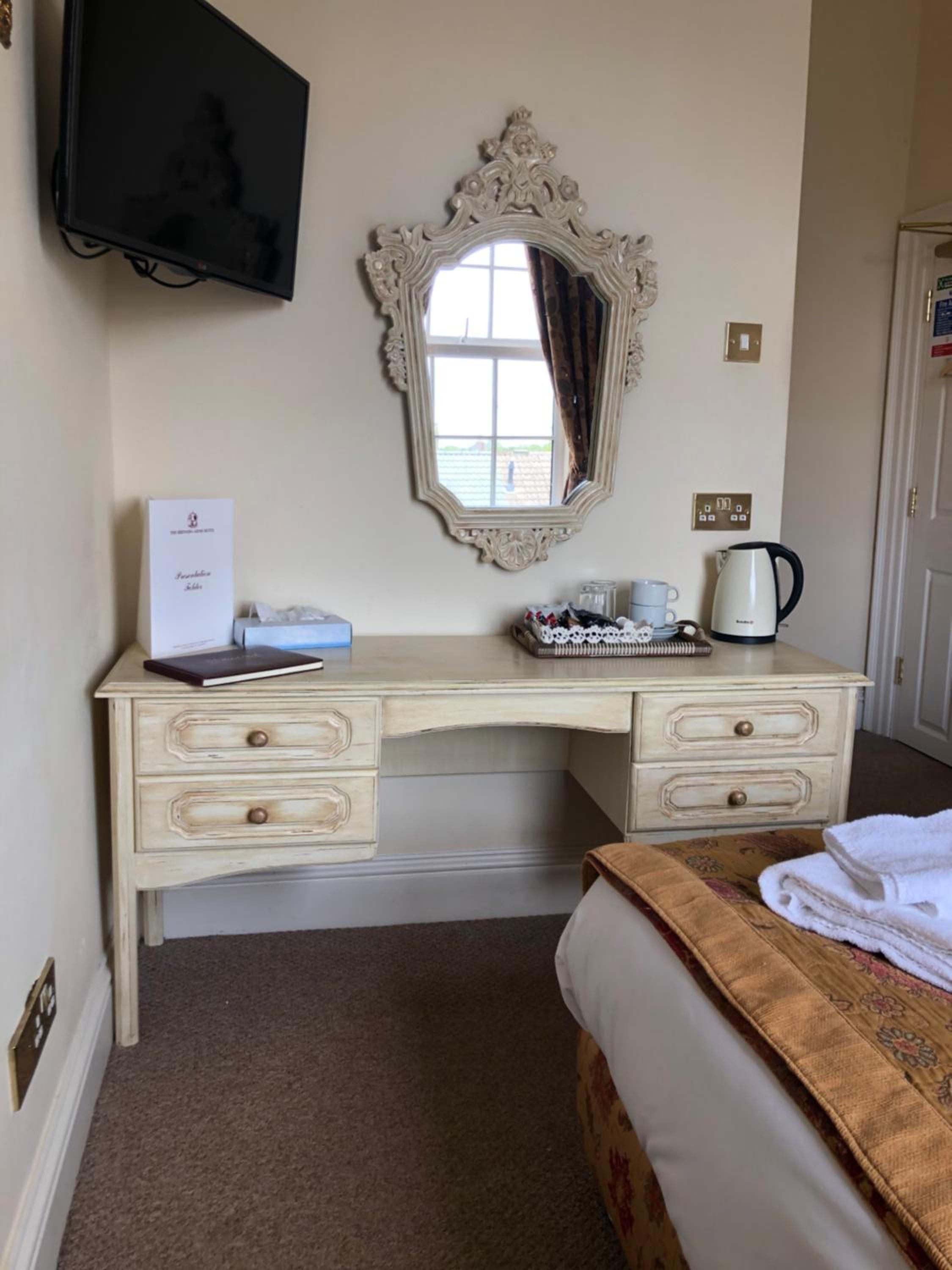 A hotel room with a bed, a desk with drawers, a television, a mirror, a window with curtains, a coffee maker, and a bedside table with a book and a tissue box.