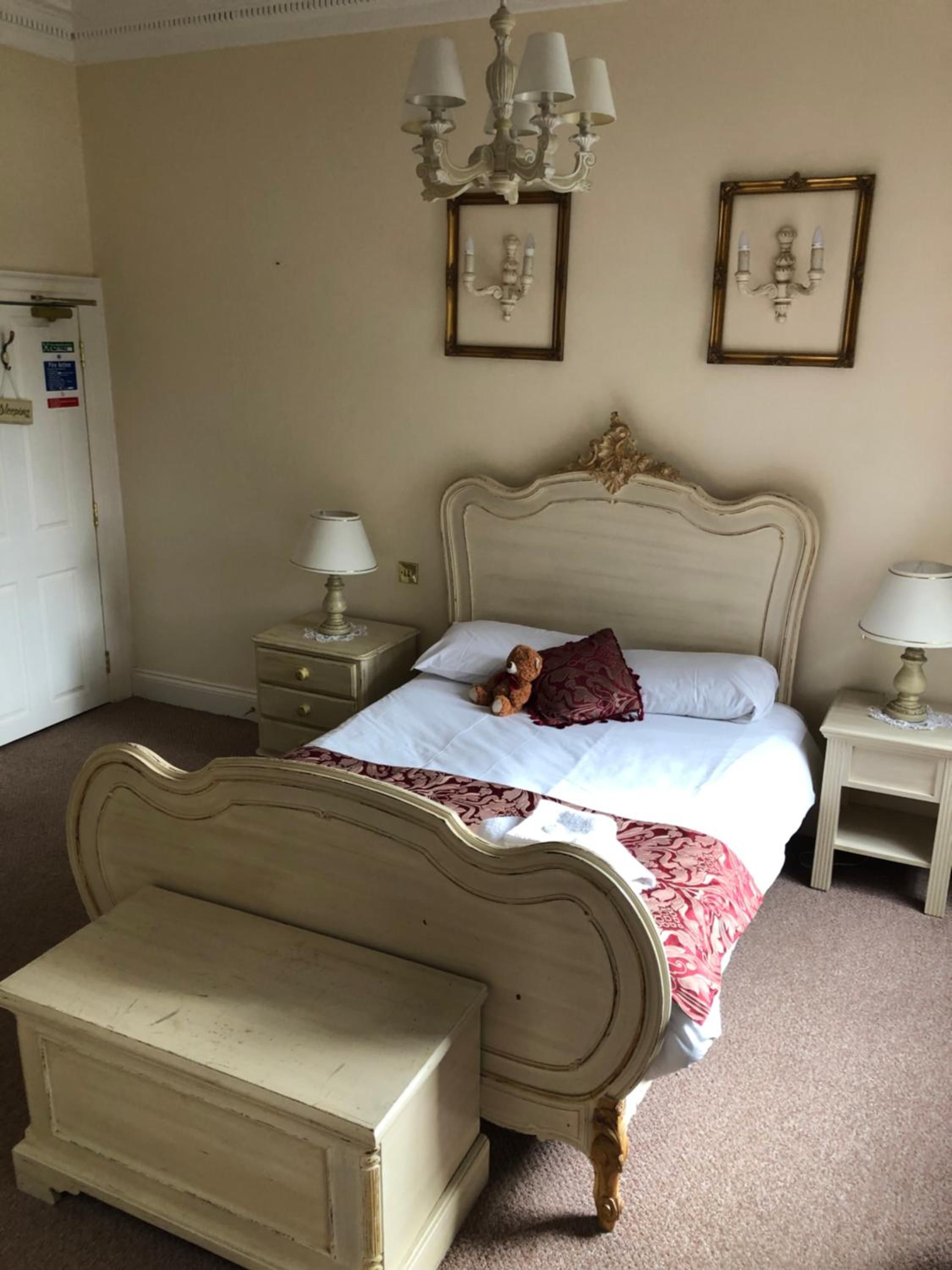 A bedroom with a bed, bedside tables, a dresser, a chandelier, and framed pictures on the wall.