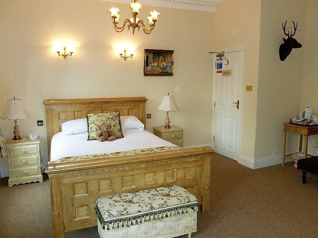 A bedroom with a wooden bed, bedside lamps, a nightstand, a bench, a wall-mounted deer head, and a door with a sign.