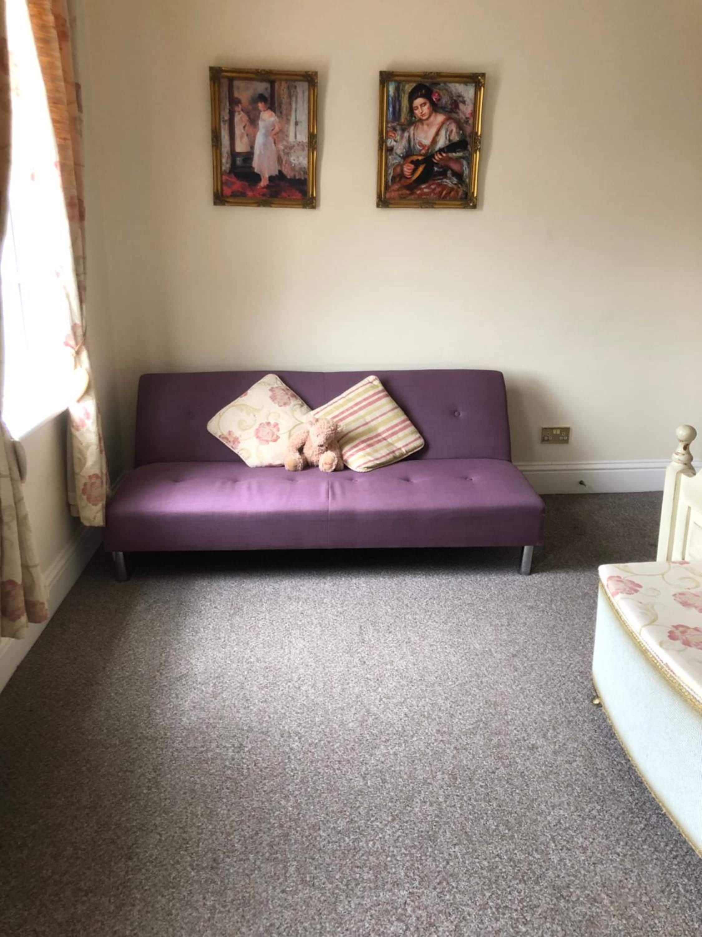 A purple sofa with floral and striped cushions, two framed paintings on the wall, and a carpeted floor.