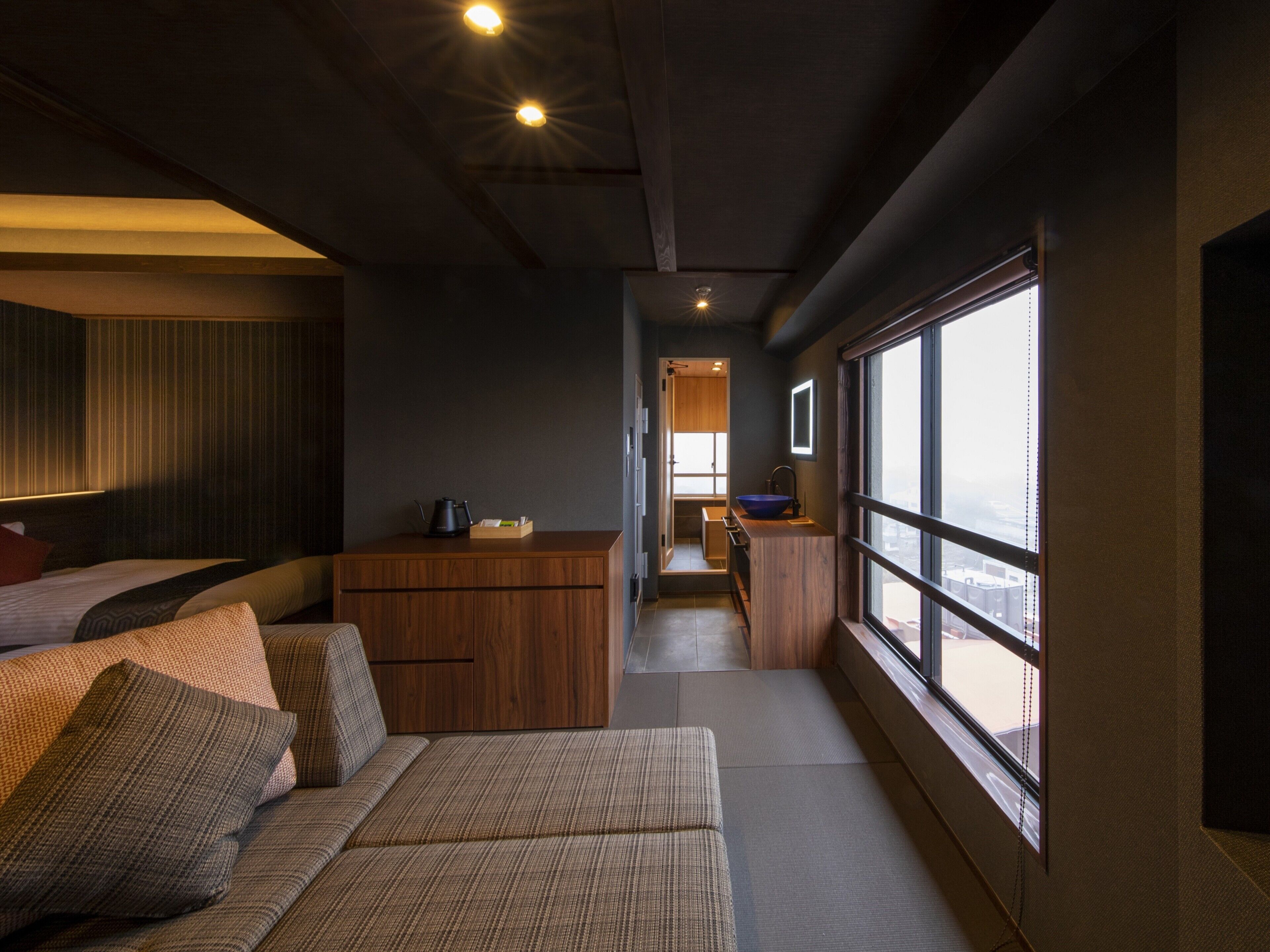 A modern hotel room with a sofa, a wooden cabinet, a small kitchen area, and a large window offering a view.